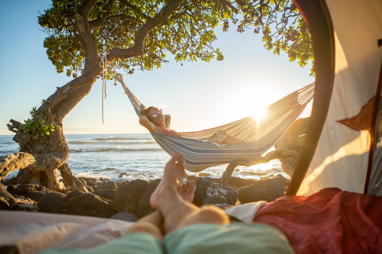 Point of view of man's feet from inside a tent camping on the beach in Hawaii looking at a woman in hammock outdoors