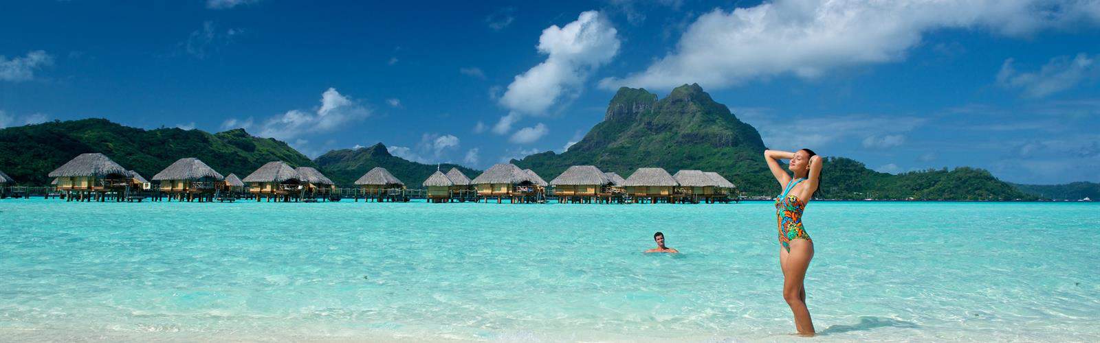 couple in tourquoise water with overwater bungalows and green tropical mountain in background
