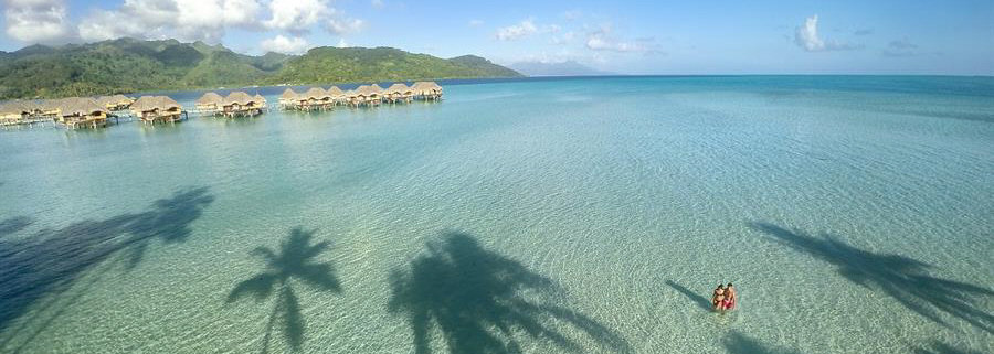 Couple wading in shallow sandy sea with overwater bungalows and island in background