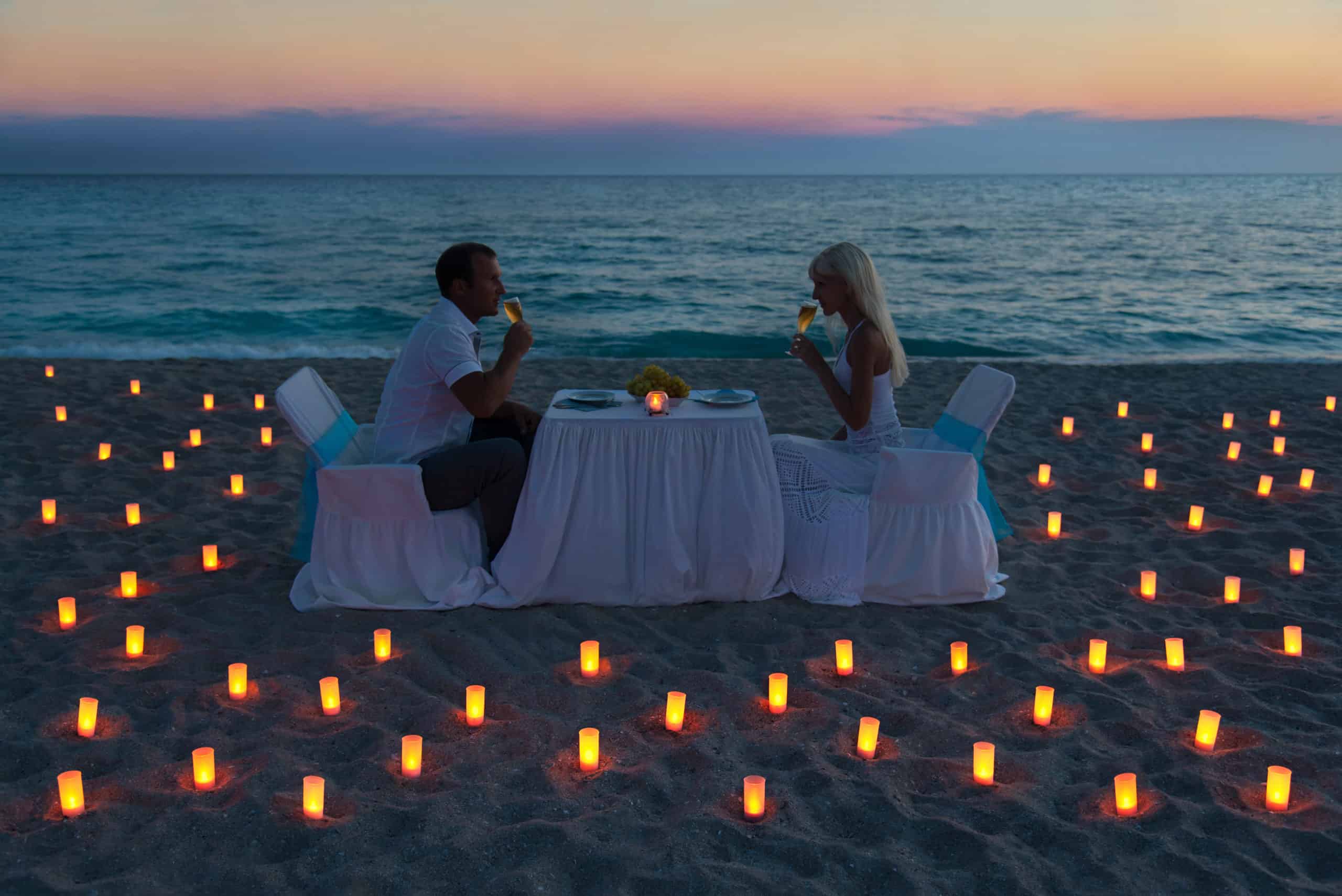 Couple drinking wine while seated at white skirted dining table on beach at sunset